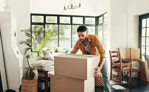 A man picking up a brown moving box that is stacked on top of another in the living room of a home.