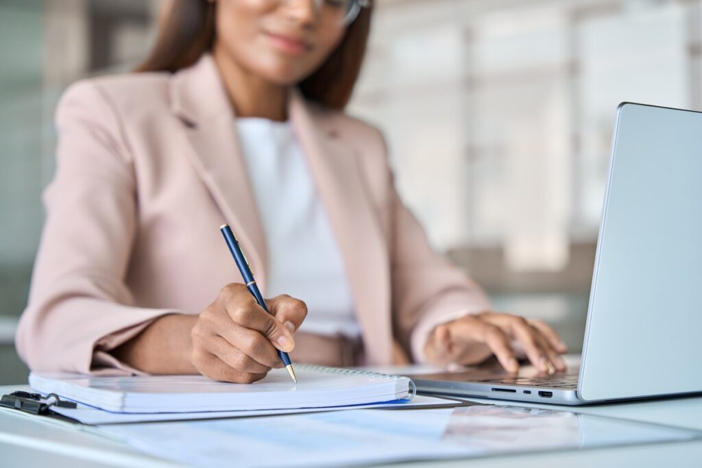 A businesswoman taking notes on a notepad while working on a laptop.