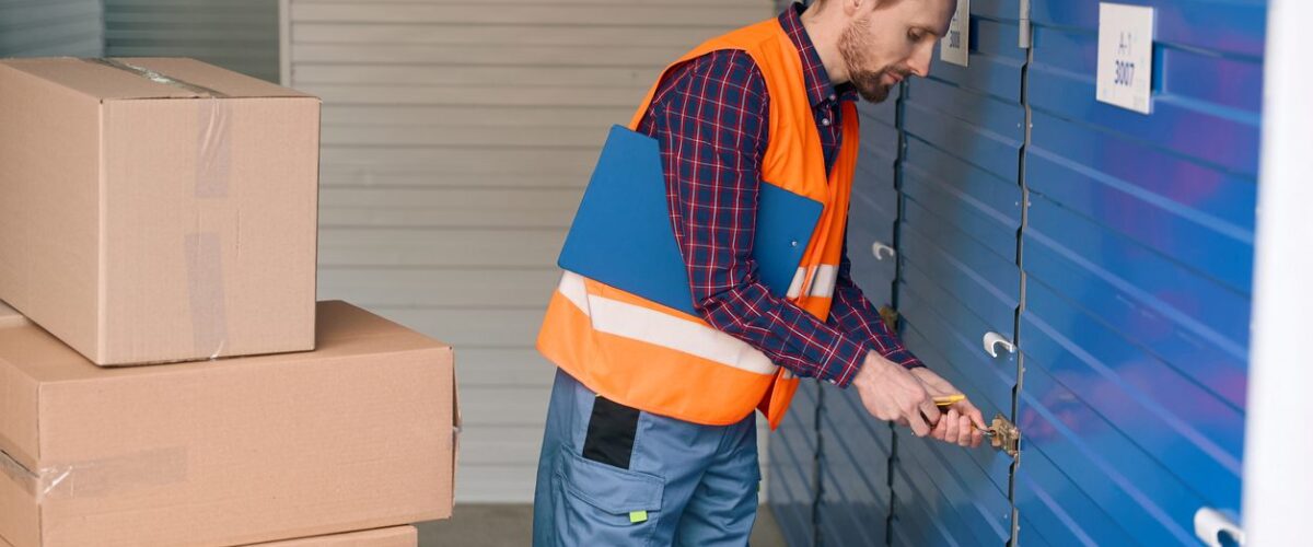 A person locking a blue storage unit door with moving boxes stacked nearby.