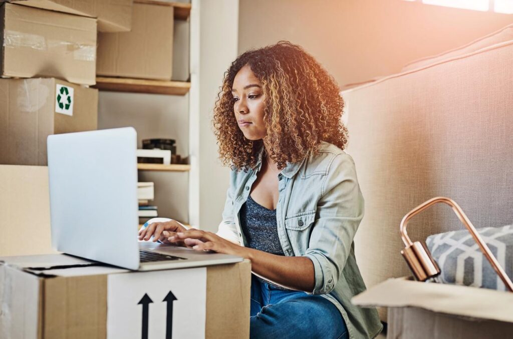 A young person using a laptop in a room filled with cardboard moving boxes.