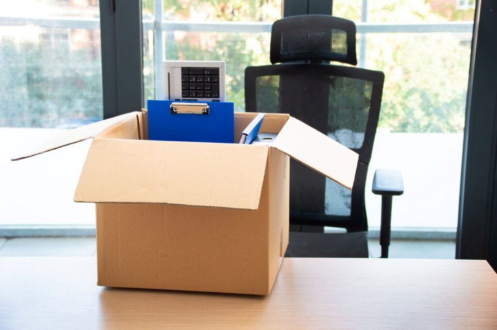 A cardboard box of office supplies sits on a desk next to an office chair.