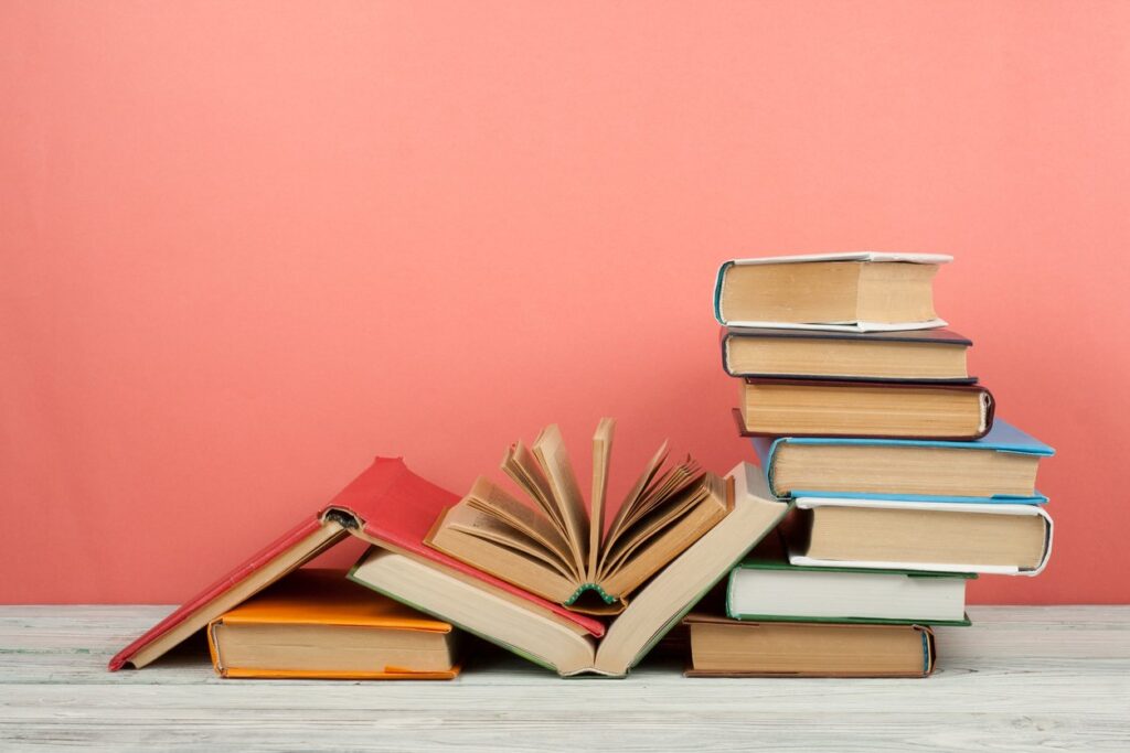Several hardcover books stacked on a wooden table in front of a pink background.
