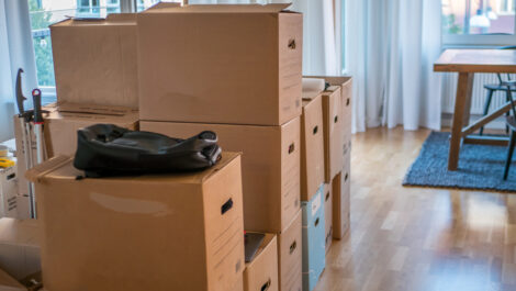 Stacks of cardboard moving boxes with an empty backpack on top at a home with wooden floors.