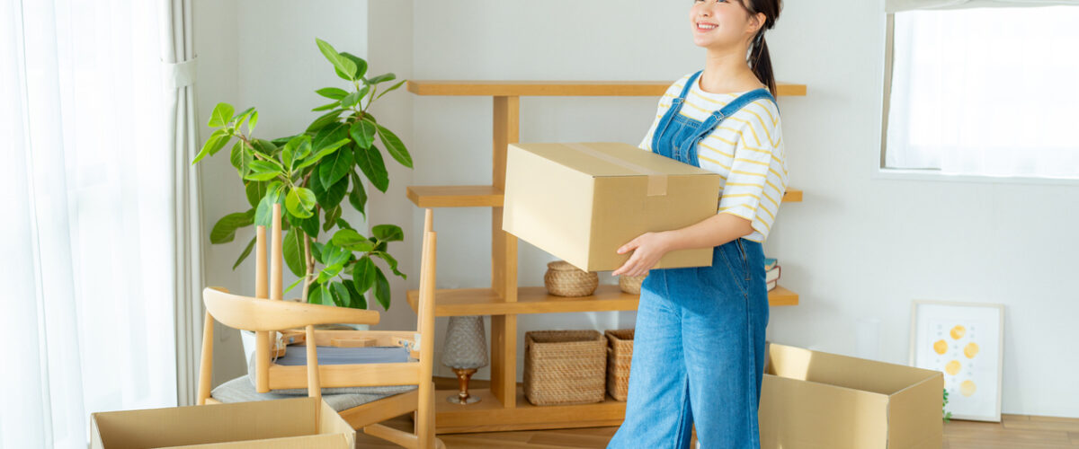 A smiling young woman carrying a cardboard box looks around her apartment, which is filled with moving boxes.