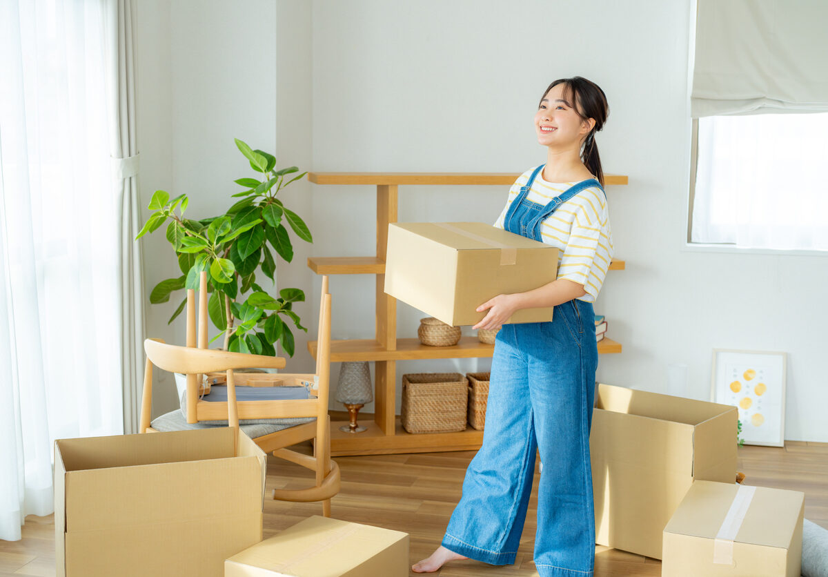 A smiling young woman carrying a cardboard box looks around her apartment, which is filled with moving boxes.