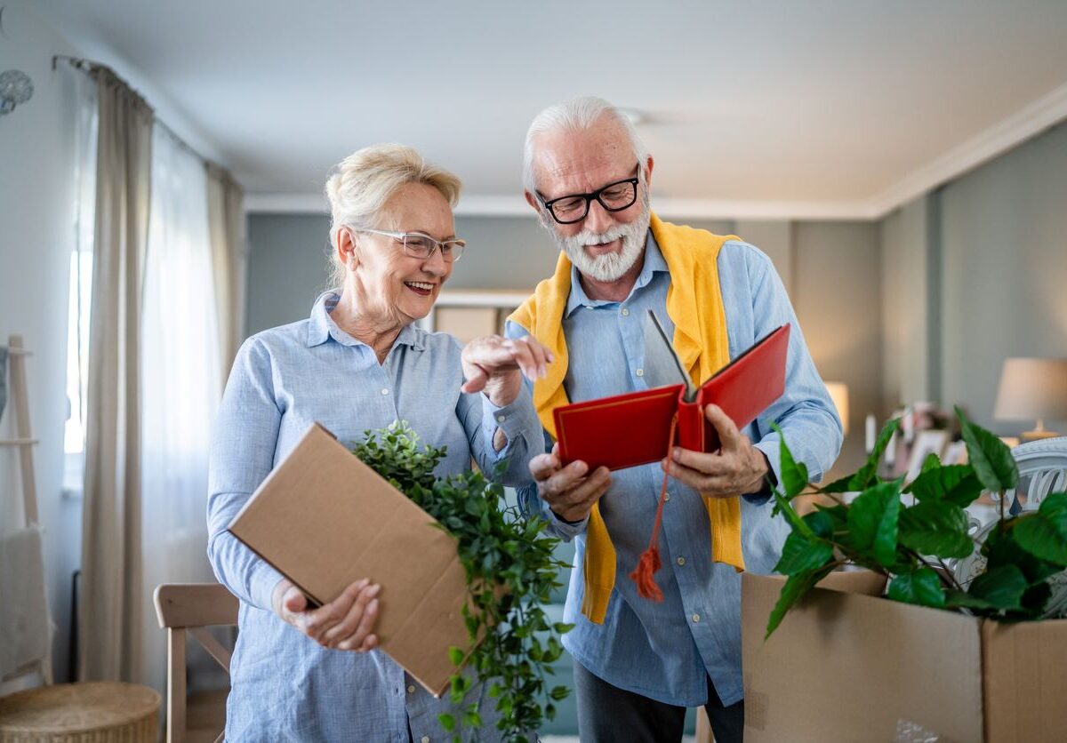 A senior couple reminisces over a photo album together as they pack cardboard boxes to move.